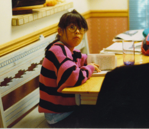 A child around ten years old, wearing a hot pink and black striped sweater and large purple glasses peeking out under her thick bangs, holding a book open at a kitchen table and looking at the camera with a surprised expression. 