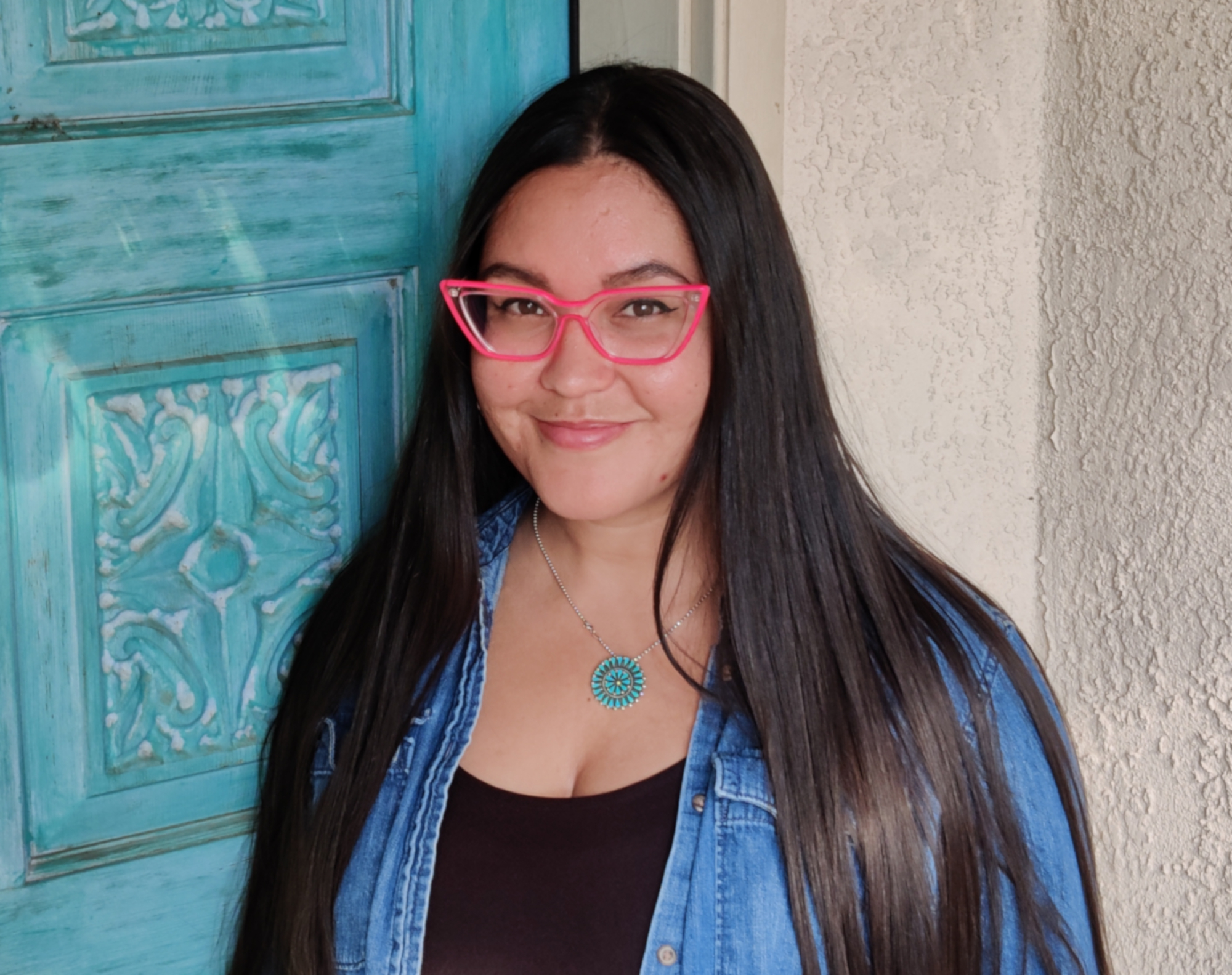 Woman with long, dark hair wearing hot pink cat eye glasses and a black top under a denim shirt, standing in front of a carved turquoise painted door and stucco wall.