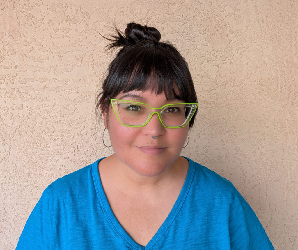Woman with dark hair up in a knot with bangs, wearing lime green cat eye glasses and a bright blue top, with sliver hoops earrings and a bright blue septum ring, standing in front of a beige stucco wall.